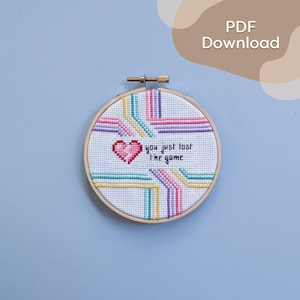 May include: A cross-stitch embroidery hoop featuring a pink pixelated heart and the text "you just lost the game." The design includes colourful diagonal lines in pink, teal, yellow, and purple. The embroidery is set in a wooden hoop.