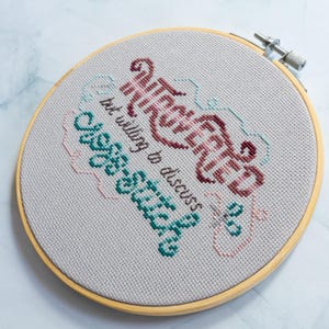 May include: A completed cross-stitch project in a wooden hoop. The text reads "Introverted but willing to discuss cross-stitch" in burgundy, teal, and pink thread on a light gray fabric. The hoop is secured with a metal clasp.