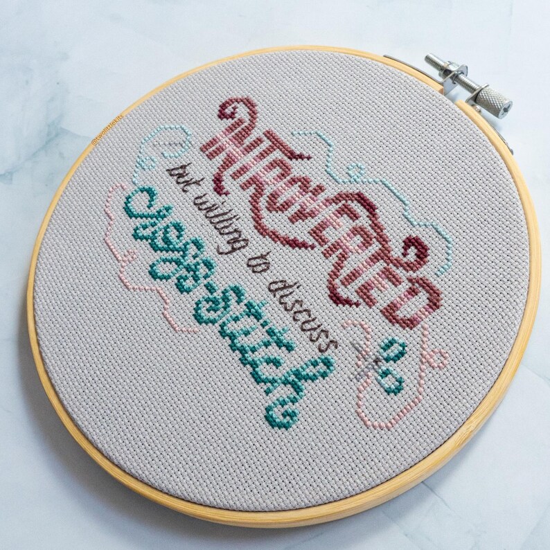 May include: A completed cross-stitch project in a wooden hoop. The text reads "Introverted but willing to discuss cross-stitch" in burgundy, teal, and pink thread on a light gray fabric. The hoop is secured with a metal clasp.
