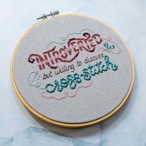 May include: A cross-stitch embroidery hoop with the words "INTROVERTED but willing to discuss cross-stitch." The text is in burgundy, teal, and pink thread on a light gray fabric, set in a wooden hoop.