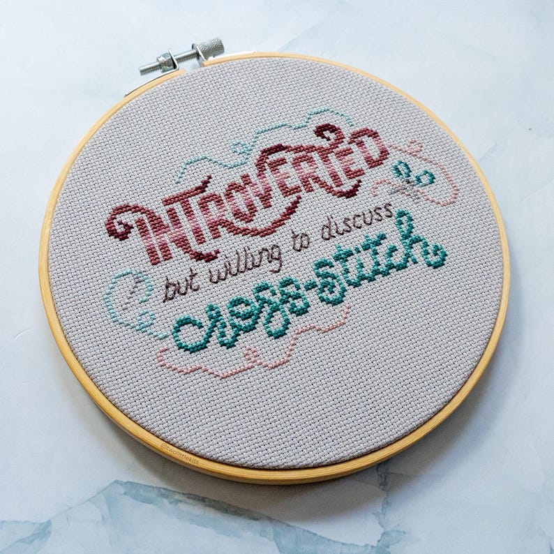 May include: A cross-stitch embroidery hoop with the words "INTROVERTED but willing to discuss cross-stitch." The text is in burgundy, teal, and pink thread on a light gray fabric, set in a wooden hoop.