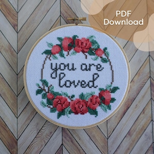 Peut inclure: Un cercle de broderie au point de croix avec les mots "you are loved" en fil noir, entouré d'une couronne de roses rouges et de feuilles vertes. La broderie est sur un tissu blanc, encadrée par un cercle en bois.