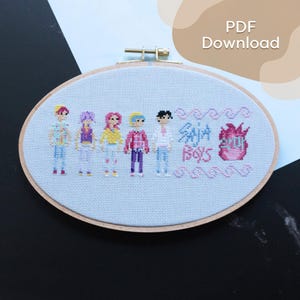 Puede incluir: Bordado de punto de cruz con cinco figuras estilizadas con atuendos coloridos, con el texto "Saja Boys" y un diseño rosa. El bordado está en un aro de madera ovalado sobre tela azul claro.