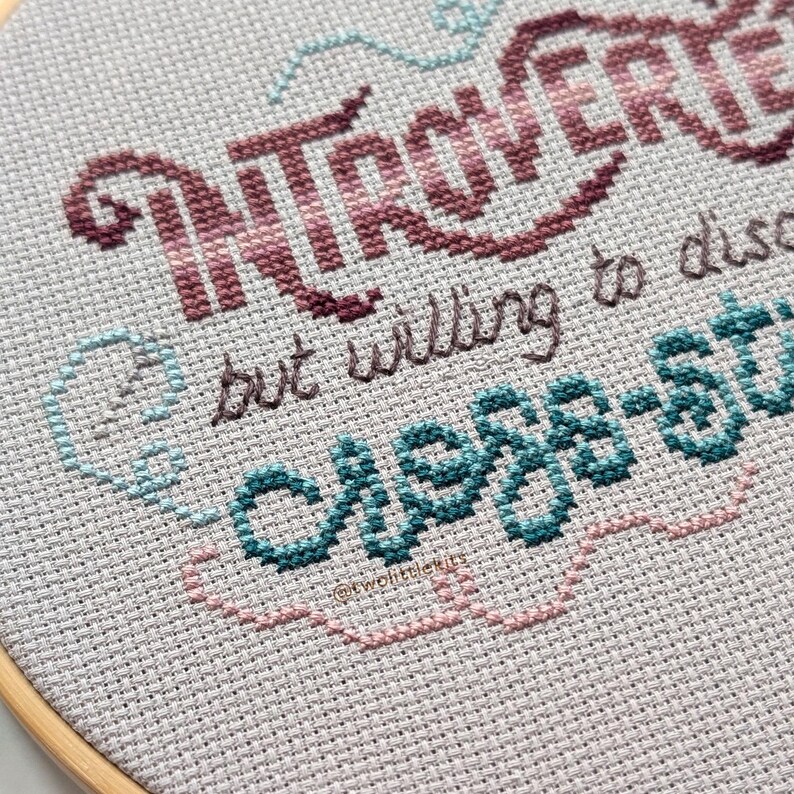May include: Cross-stitch embroidery in a wooden hoop features the word "INTROVERT" in burgundy and pink, with the text "but willing to discuss" and "chess-stuff" in teal and brown. The design is on a light gray fabric.