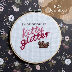 Puede incluir: Un aro de bordado blanco con un diseño de punto de cruz que dice "It's not cat hair, it's kitty glitter" en letras rosa y rojas. Un pequeño gato marrón está bordado debajo del texto.