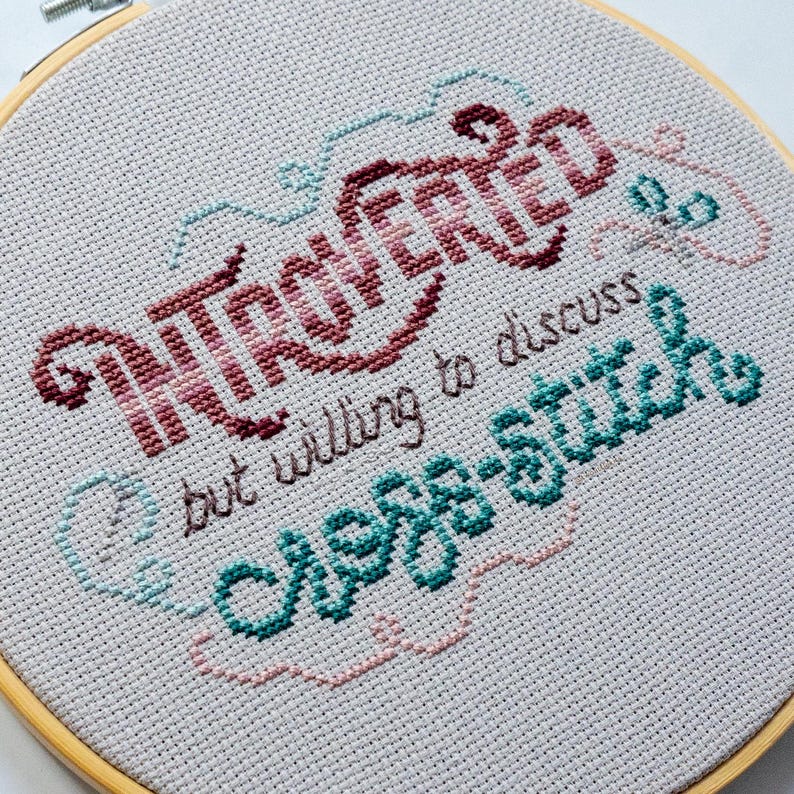 May include: Cross-stitch embroidery in a wooden hoop. The text reads "Introverted but willing to discuss cross-stitch" in burgundy, teal, and light blue thread on a light gray fabric. The design includes decorative swirls.