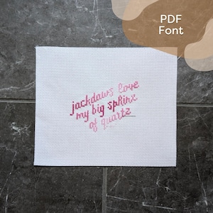 May include: A white cross-stitch fabric with the text "jackdaws love my big sphinx of quartz" in pink thread. The fabric is rectangular and set against a dark gray background. The words are in a handwritten style.