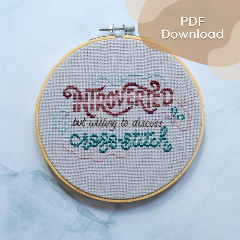 May include: Cross-stitch embroidery in a wooden hoop. The text reads "Introverted but willing to discuss cross-stitch" in burgundy, teal, and pink thread. The background is a light gray fabric.