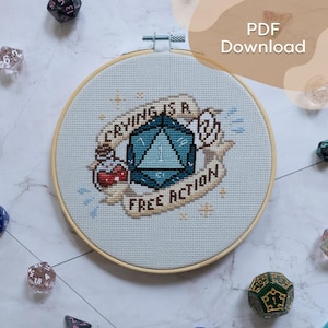 Pode incluir: Um bastidor de bordado circular de madeira contendo um design em ponto cruz sobre tecido claro. O design apresenta um dado azul de vinte lados, um pequeno frasco de poção com líquido vermelho e uma faixa com o texto "CRYING IS A FREE ACTION".