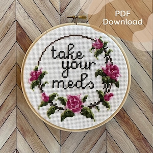 May include: A white embroidery hoop with a cross-stitch design featuring a floral wreath with pink roses and green leaves surrounding the text "take your meds".
