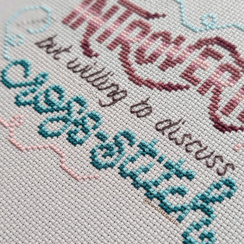 May include: Close-up of a cross-stitch design on a light gray fabric. The text reads "INTROVERT but willing to discuss SEWING" in shades of teal, pink, and burgundy. The design is framed with a light blue and pink border.