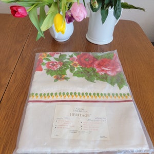 May include: A white tablecloth with a red rose floral pattern. The tablecloth is packaged in plastic and has a label that reads "Nappe Tablecloth Heritage Rose 60x84" inches.