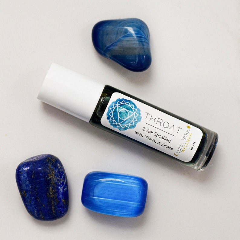 May include: A blue glass roller bottle with a label that reads "Throat - I Am Speaking with Truth & Grace - CLUNA SOUL WELLNESS - 10 mL". The bottle is on a white surface with three blue stones.