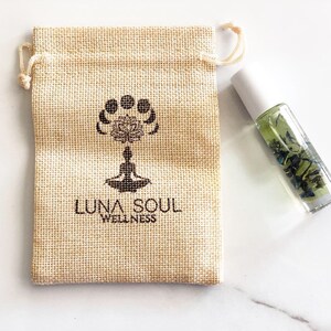 May include: A small, beige hessian pouch with a drawstring closure. The pouch has a black logo that says "Luna Soul Wellness" and a design of a lotus flower with moons around it. A clear glass bottle with a white cap and green liquid inside is next to the pouch.