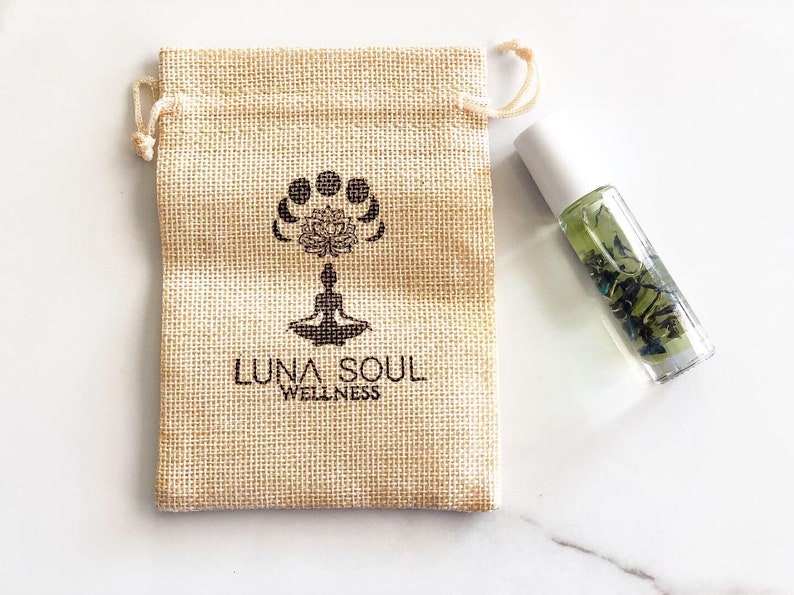May include: A small, beige hessian pouch with a drawstring closure. The pouch has a black logo that says "Luna Soul Wellness" and a design of a lotus flower with moons around it. A clear glass bottle with a white cap and green liquid inside is next to the pouch.