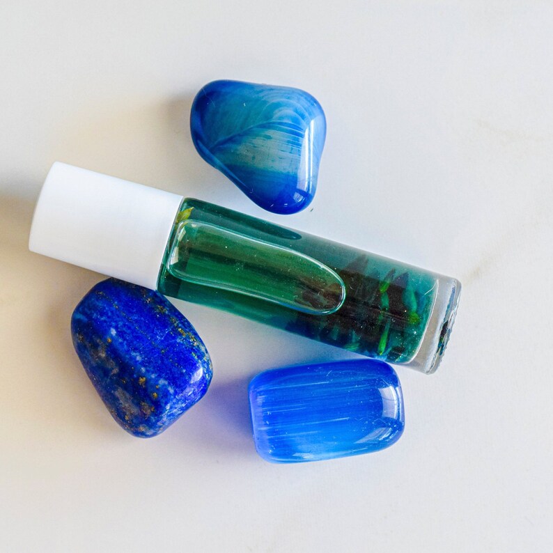 May include: A green glass roller bottle with a white cap, next to three blue gemstones. The gemstones are various shades of blue and have a smooth, polished finish.