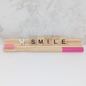 May include: A wooden toothbrush with a pink handle and bristles, paired with a wooden holder displaying the word "SMILE" in Scrabble tiles. A white tooth-shaped decoration is also present.