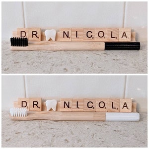 May include: Two wooden toothbrushes with black and white bristles, a white tooth-shaped tile, and a set of wooden tiles spelling "DR. NICOLA" on a light brown surface.