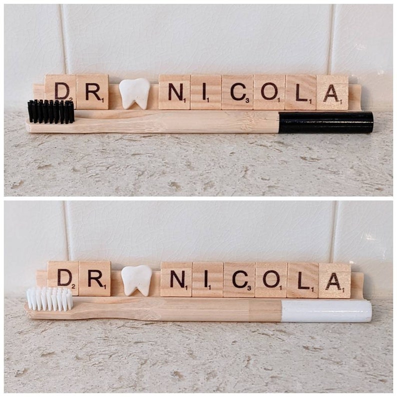 May include: Two wooden toothbrushes with black and white bristles, a white tooth-shaped tile, and a set of wooden tiles spelling "DR. NICOLA" on a light brown surface.