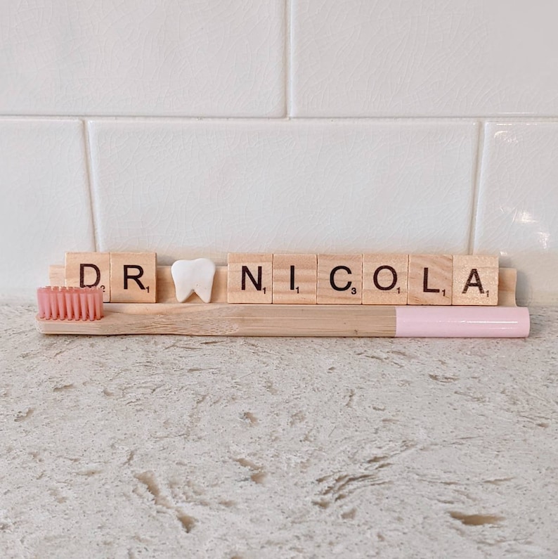May include: Wooden tiles spelling "DR. NICOLA" with a white tooth tile in between the "R" and "N". A pink and wooden toothbrush is in front of the tiles.