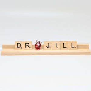 Può includere: La parola "DR. JILL" è scritta con tessere di lettere su un supporto in legno. Un ciondolo a forma di cuore rosso è posizionato tra la "R" e la "J".