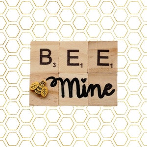 Può includere: Tessere di lettere in legno che compongono la parola "BEE" con un piccolo ciondolo a forma di ape gialla e la parola "Mine" scritta in nero su un blocco di legno. Lo sfondo è un motivo a nido d'ape dorato.
