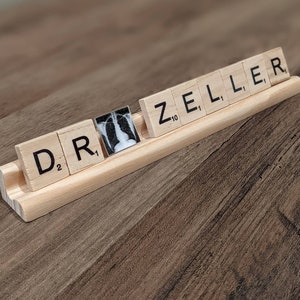 Può includere: Un rack di legno con la parola "DR" e una tessera vuota seguita dalla parola "ZELLER" scritta su tessere nere. Le tessere sono su una superficie di legno.