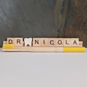 May include: A wooden tile spelling "DR. NICOLA" with a white tooth charm in between the "R" and "N" tiles. A yellow toothbrush is in front of the tiles.