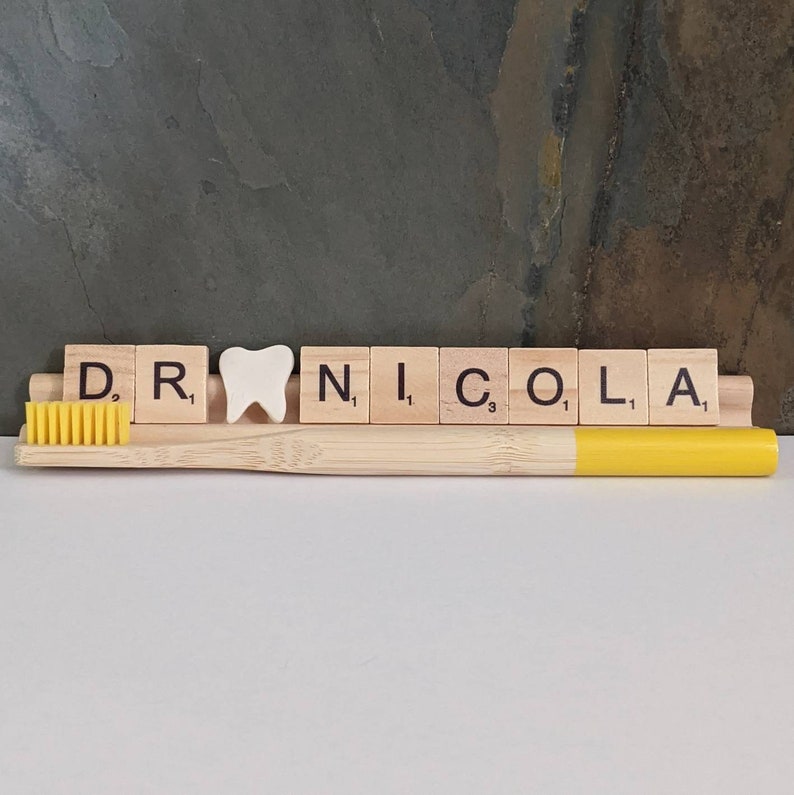 May include: A wooden tile spelling "DR. NICOLA" with a white tooth charm in between the "R" and "N" tiles. A yellow toothbrush is in front of the tiles.