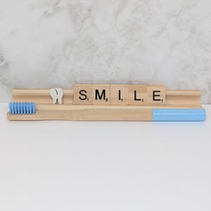 May include: A bamboo toothbrush with a light blue handle and bristles, next to a wooden holder with the word "SMILE" spelled out in letter tiles and a tooth-shaped decoration. The toothbrush is a dental hygiene product.