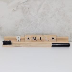 May include: A bamboo toothbrush with black bristles and a black handle, next to a wooden holder with the word "SMILE" spelled out in letter tiles. A white tooth-shaped decoration is also present.