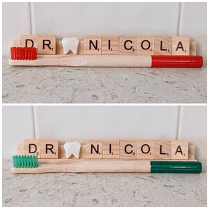 May include: Two wooden toothbrushes with red and green bristles. The toothbrushes are next to a set of letter tiles spelling out "DR. NICOLA".