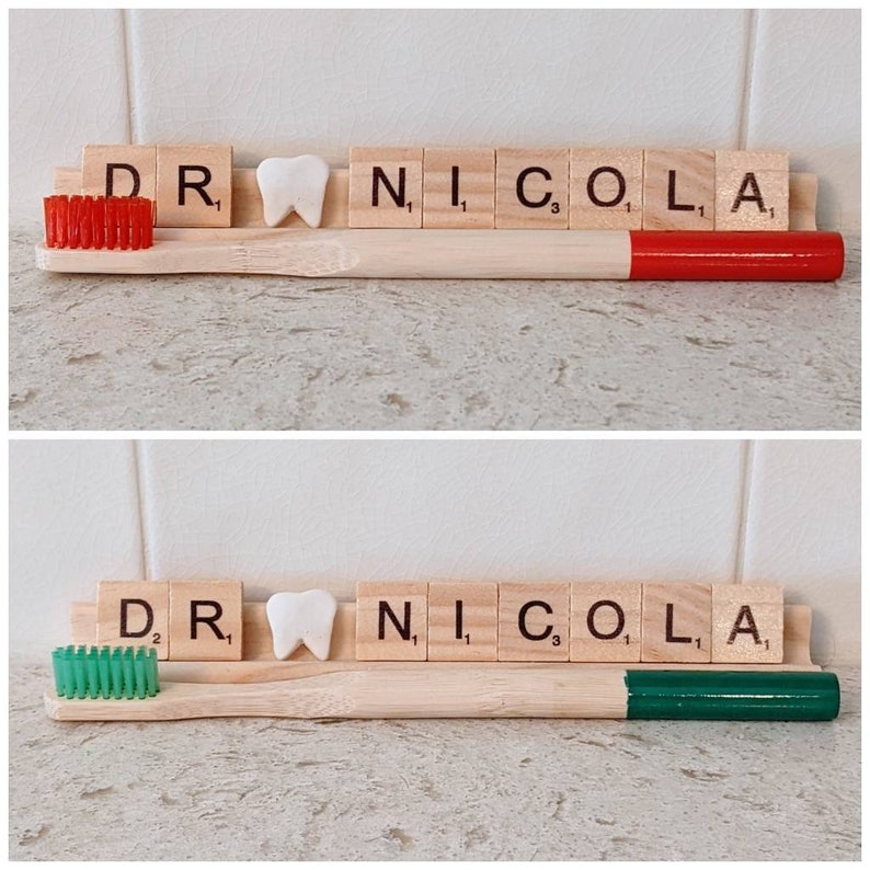May include: Two wooden toothbrushes with red and green bristles. The toothbrushes are next to a set of letter tiles spelling out "DR. NICOLA".