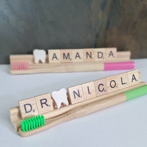 May include: Two wooden toothbrush holders with the names "Amanda" and "Nicola" spelled out in white tiles. Each holder has a white tooth model and a green and pink toothbrush.