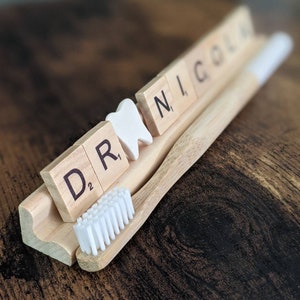 May include: A wooden tray with the words "DR", "N", "I", and "COL" spelled out in white letters on wooden tiles. A white tooth-shaped object is placed between the "DR" and "N" tiles. A bamboo toothbrush with white bristles is placed on the tray.