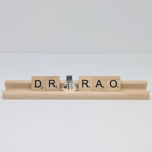 May include: A wooden tile holder with Scrabble tiles spelling "DR RAO". A small, silver-colored flask is placed between the letters. The tiles and holder are made of light-colored wood, set against a white background.