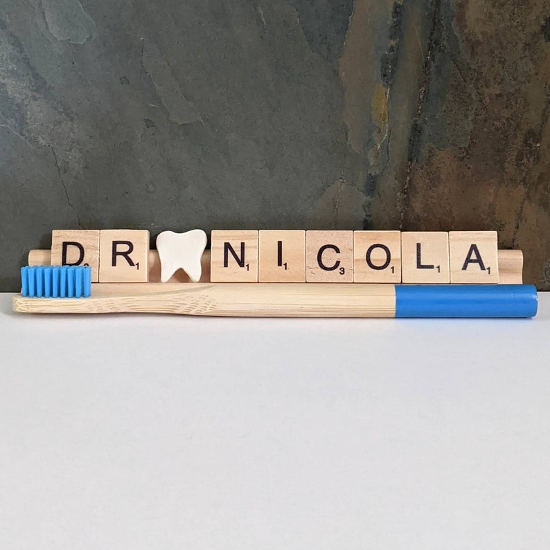 May include: Wooden letter tiles spelling "DR. NICOLA" with a white tooth charm in between the "R" and "N". A blue and wooden toothbrush is in front of the tiles.