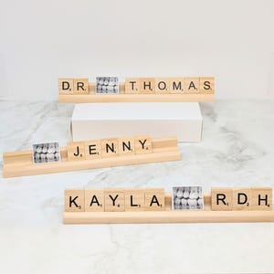 May include: Three wooden name plaques with the names "DR. THOMAS", "JENNY", and "KAYLA, RDH" spelled out in black letter tiles. Each plaque has a small black and white x-ray image of teeth.