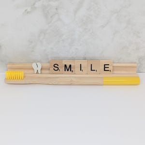 May include: A bamboo toothbrush with a yellow handle and bristles, and a wooden holder with the word "SMILE" spelled out in Scrabble tiles. A white tooth-shaped decoration is also present.