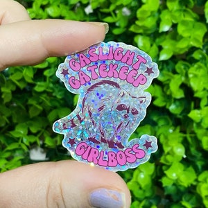 Peut inclure: Un autocollant holographique avec une illustration de raton laveur et le texte "GASLIGHT GATEKEEP GIRLBOSS" en rose et blanc. L'autocollant est tenu devant un fond de feuillage vert.