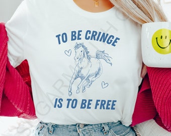 May include: White t-shirt with a blue illustration of a horse and the text "TO BE CRINGE IS TO BE FREE." A white mug with a yellow smiley face is held in the hand. The person is wearing blue jeans and a pink cardigan.