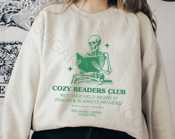 May include: A cream-coloured sweatshirt featuring a green graphic of a skeleton reading a book. The text "COZY READERS CLUB" is printed below the graphic, along with details about meetings, snacks, blankets, and the genre "romantasy".