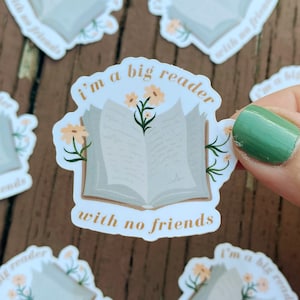 May include: A sticker with the text "I'm a big reader with no friends" in gold lettering. The sticker features an open book design with flowers and leaves. Several stickers are scattered on a wooden surface.