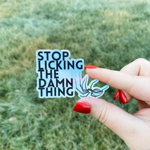 May include: A holographic sticker with the text "STOP LICKING THE DAMN THING" in black, along with a black and white spider illustration. The sticker is held between fingers with red nail polish, set against a blurred green background.
