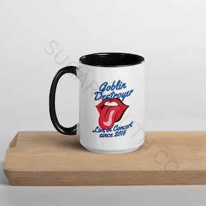 May include: White ceramic mug with a black handle and interior. The mug features the text "Goblin Destroyer" in blue, a red tongue graphic, and the text "Live in Concert since 2016". The mug sits on a wooden surface.