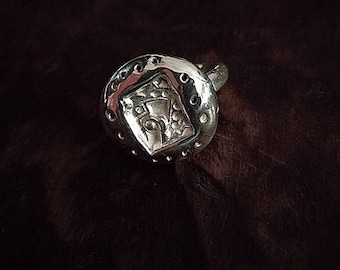 May include: A silver-colored bottle stopper with a round shape and a decorative design. The stopper features a raised, square-shaped emblem with an ornate pattern and is surrounded by small, evenly spaced holes.