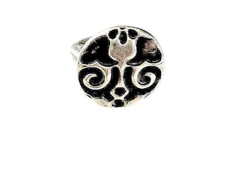May include: A silver ring with a circular face featuring a black and silver abstract design. The design includes a central shape with curved and swirling elements. The ring is set against a white background.