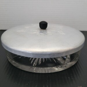 May include: A round, clear glass container with a decorative, cut-glass design. It has a silver-colored metal lid with a black knob. The lid has a floral design etched into the metal.