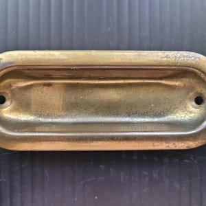 May include: A vintage brass recessed pull with an oval shape. The pull has a rectangular indentation and two small holes for mounting. The brass has a worn, aged appearance.