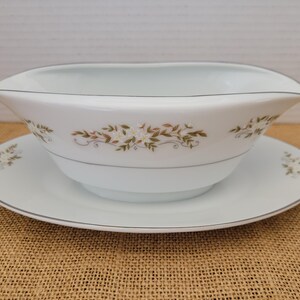 May include: A white porcelain gravy boat and saucer set. The boat has a spout and is decorated with a floral pattern in gold and green. A thin silver-coloured line trims the edges of both pieces. The saucer is round.